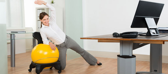 Frau macht aktive Pause im Büro. Streckt Gliedmaßen vor Schreibtisch mit Hilfe eines gelben Gymnastikballs.