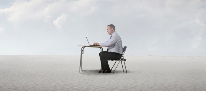 Ein Mann sitzt am Schreibtisch, mitten in der Wüste, und arbeitet an seinem Laptop. Weit und breit ist nichts um ihn herum zu sehen, außer Sand.