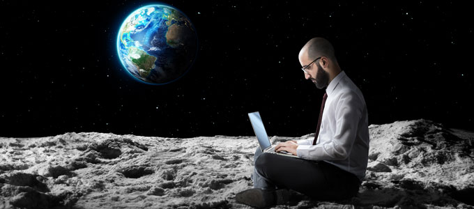 Ein Mann sitzt im Schneidersitz mit seinem Laptop auf den Beinen im Weltall, höchstwahrscheinlich auf dem Mond. Im Hintergrund ist das schwarze Weltall mit vereinzelten Sternen und die von der Sonne angestrahlte Erde zu sehen.