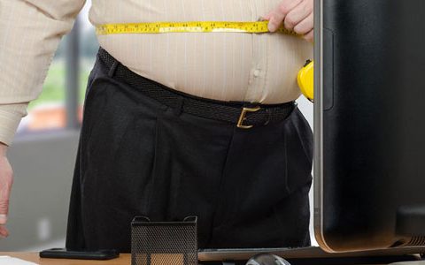 Ein übergewichtiger Mann steht am Arbeitsplatz und misst seinen Bauch mit einem Maßband.