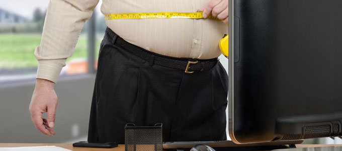 Ein übergewichtiger Mann steht am Arbeitsplatz und misst seinen Bauch mit einem Maßband.