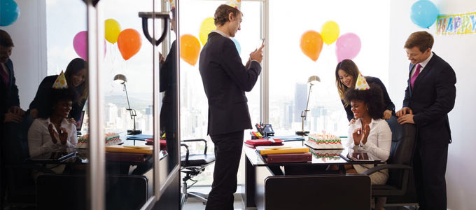 Ein Büro, geschmückt mit bunten Luftballons und Happy Birthday Banner. Inmitten der Räumlichkeiten sind vier Personen am Schreibtisch versammelt. Eine junge Frau mit Partyhut auf dem Kopf sitzt auf dem Bürostuhl und strahlt vor Freude. Vor ihr auf dem Tisch eine Geburstagstorte. Um sie herum stehen zwei Kollegen und eine Kollegin. Einer der Kollegen fotografiert das Szenario mit seinem Smartphone.
