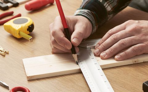 Handwerk als Hobby / auf dem Foto: Ein Mann bereitet ein DIY-Projekt vor und misst ein Holzstück aus. Handwerker malt mit Hilfe eines Lineals mit einem Bleistift eine Linie auf ein Holzbrett. Um ihn herum liegen weitere Handwerksutensilien wie Nägel, Maßband, Zange, Hammer und Schraubendreher. Fokus des Bildes sind die Hände des Handwerkers.