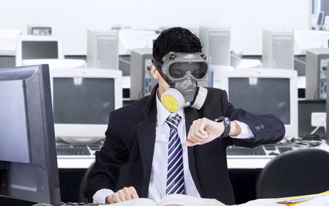 Luftqualität im Büro / auf dem Foto: Ein Angestellter sitzt mit Gasmaske in einem Büroraum voller Computer und schaut auf die Uhr. Geschäftsmann, in Anzug und Krawatte, sitzt am Arbeitsplatz im Großraumbüro, umgeben von zahleichen Computern und Bildschirmen, mit Gasmaske und Augenschutzbrille und schaut auf seine Armbanduhr.
