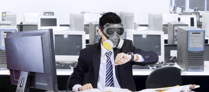 Luftqualität im Büro / auf dem Foto: Ein Angestellter sitzt mit Gasmaske in einem Büroraum voller Computer und schaut auf die Uhr. Geschäftsmann, in Anzug und Krawatte, sitzt am Arbeitsplatz im Großraumbüro, umgeben von zahleichen Computern und Bildschirmen, mit Gasmaske und Augenschutzbrille und schaut auf seine Armbanduhr.