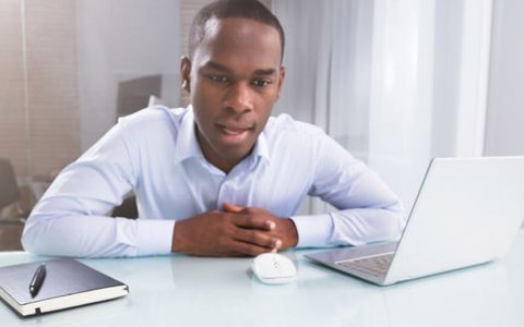 Digitale Spracherkennung / auf dem Foto: Geschäftsmann diktiert der Software einen Text. Ein Mann sitzt an einem Schreibtisch, sein Büro ist sehr schlicht und in weiß gehalten. Er selber trägt ein weißes Hemd. Er spricht offensichtlich mit einem Sprachassistenten, den er dabei mit den Augen fixiert.