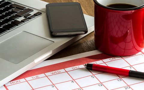 Welcher Kalender-Typ sind Sie? / auf dem Foto: Monatskalender aus Papier liegt neben Smartphone und Notebook auf dem Schreibtisch. Schreibtisch mit folgenden Utensilien: Laptop, Smartphone, rote Kaffeetasse mit schwarzem Kaffee gefüllt, Terminplaner und Kugelschreiber.