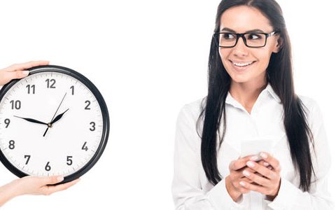 Brückenteilzeitgesetz - fröhliche Frau schaut auf eine große Uhr, die von jemanden ins Bild gehalten wird. Weißer Hintergrund. Die Frau ist weiß gekleidet und hat schwarze Haare. Die Uhr hat ein wießes Zifferblatt schwarze Zeiger und einen schwarzen Rand.