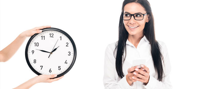 Brückenteilzeitgesetz - fröhliche Frau schaut auf eine große Uhr, die von jemanden ins Bild gehalten wird. Weißer Hintergrund. Die Frau ist weiß gekleidet und hat schwarze Haare. Die Uhr hat ein wießes Zifferblatt schwarze Zeiger und einen schwarzen Rand.