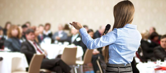 Checkliste für das perfekte Tagungshotel / auf dem Foto: Geschäftsfrau hält einen Vortrag auf einer Konferenz mit vielen Teilnehmern.