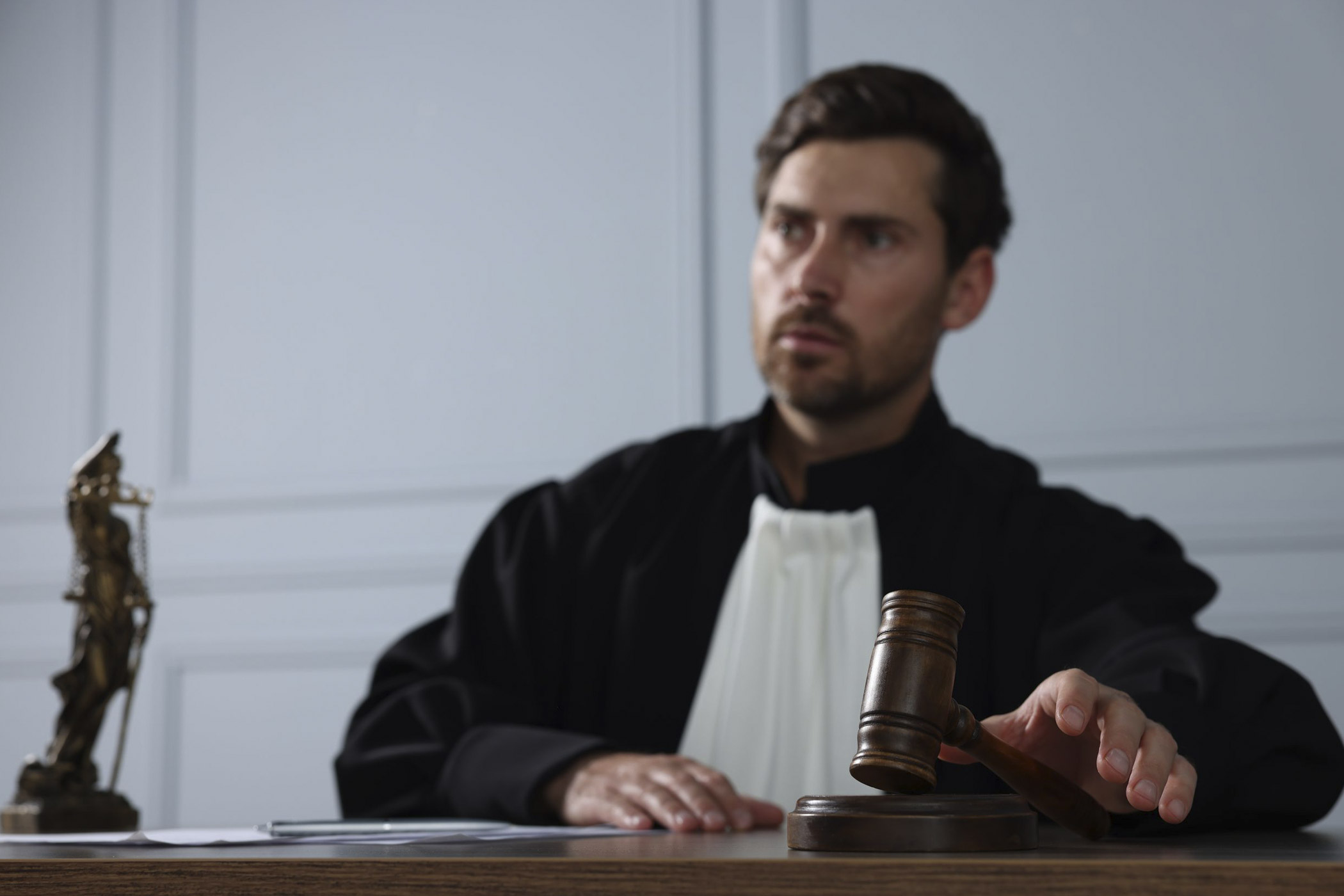 Ein Richter in schwarzer Robe mit weißem Kragen sitzt an einem Holztisch und hebt eine hölzerne Richterglocke. Im Vordergrund liegen Dokumente und ein Tablet, während im Hintergrund eine Justitia-Statue zu sehen ist. Die Szene wirkt förmlich und konzentriert.