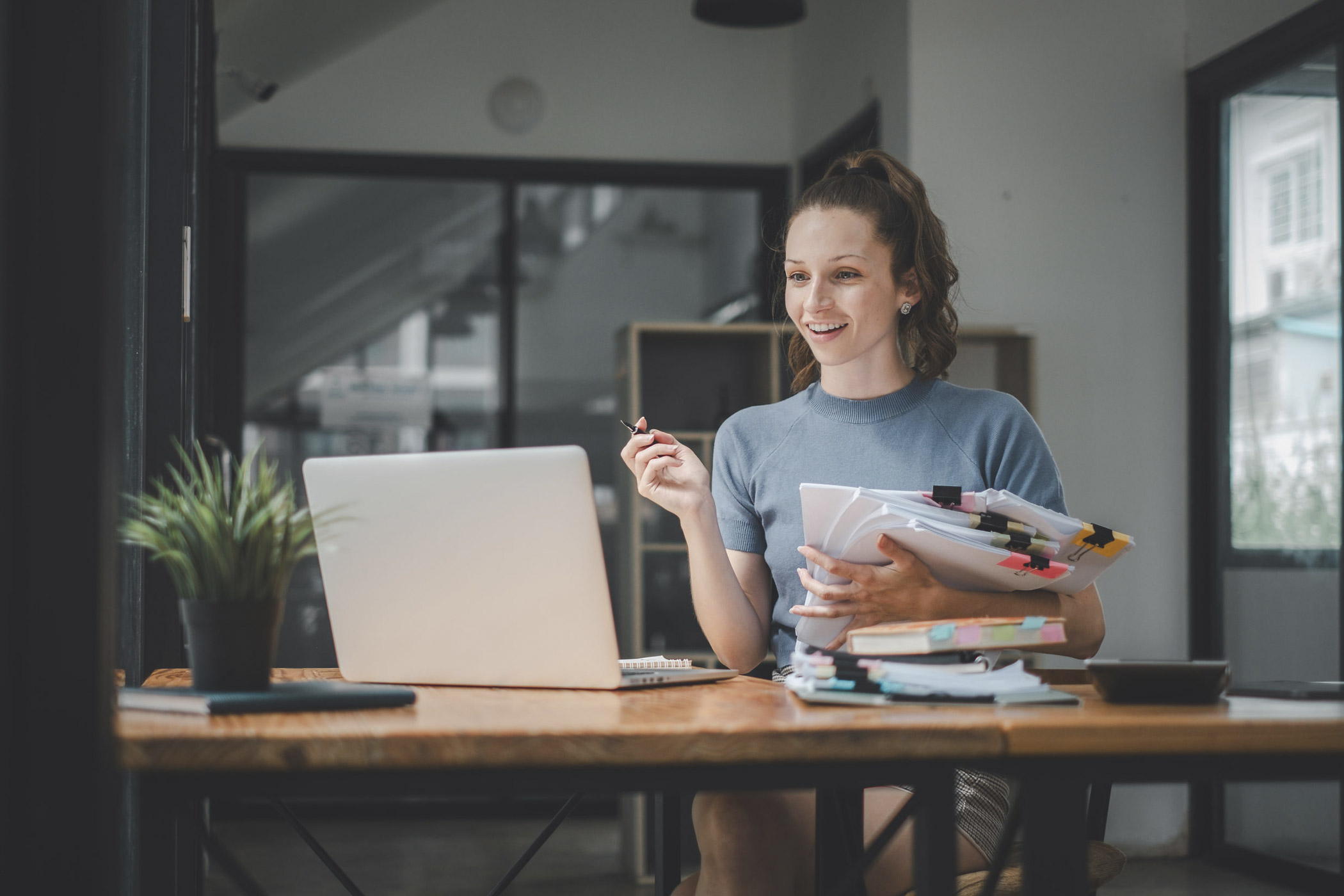 der Spagat zwischen zwei Jobs / auf dem Foto: Junge Arbeitnehmerin sitzt vor dem Laptop und hält mehrere Papierdokumente im Arm.