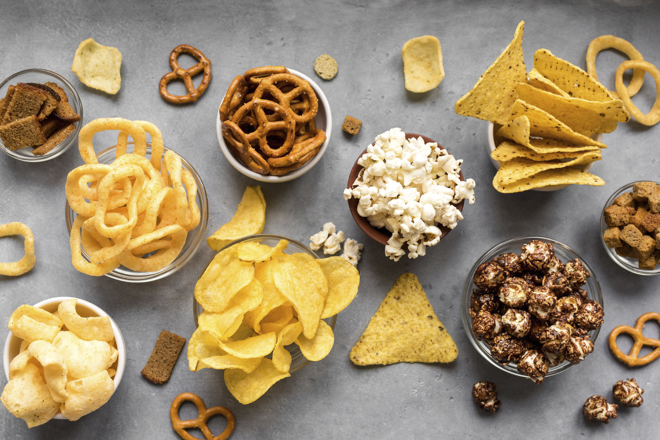 Verschiedene Schalen mit herzhaften und süßen Snacks wie Chips, Popcorn, Salzbrezeln, Nachos und Knabbergebäck auf grauer Tischoberfläche – von oben fotografiert.