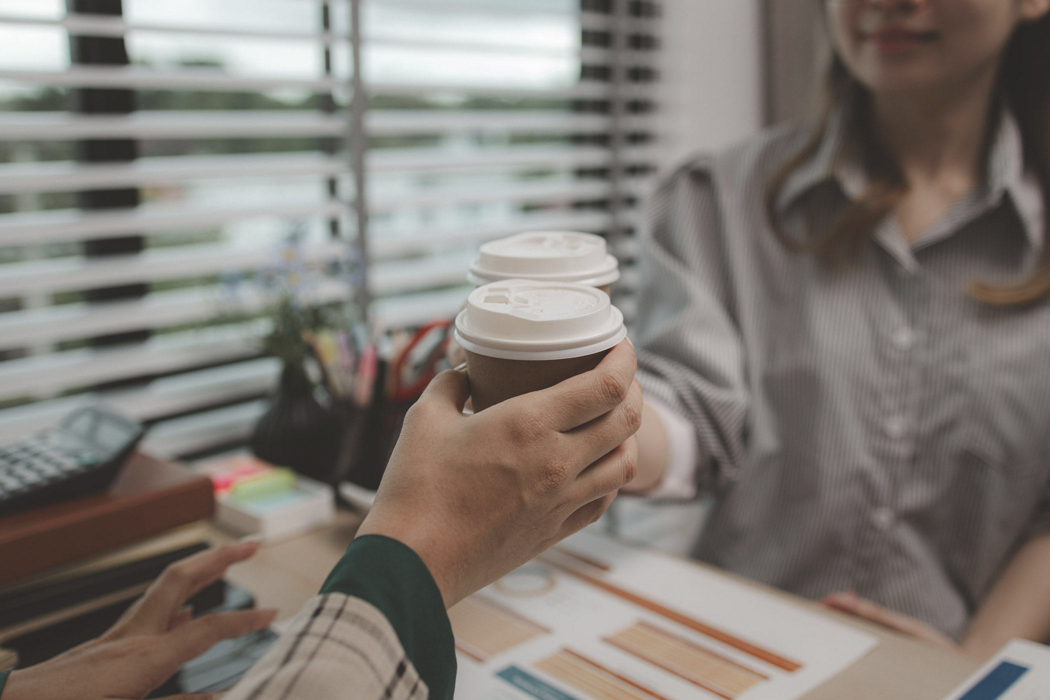 Zwei Personen im Büro stoßen mit Kaffeebechern zum Anstoßen an. Im Hintergrund ist ein Arbeitsplatz mit Büromaterialien und einem Laptop zu sehen.