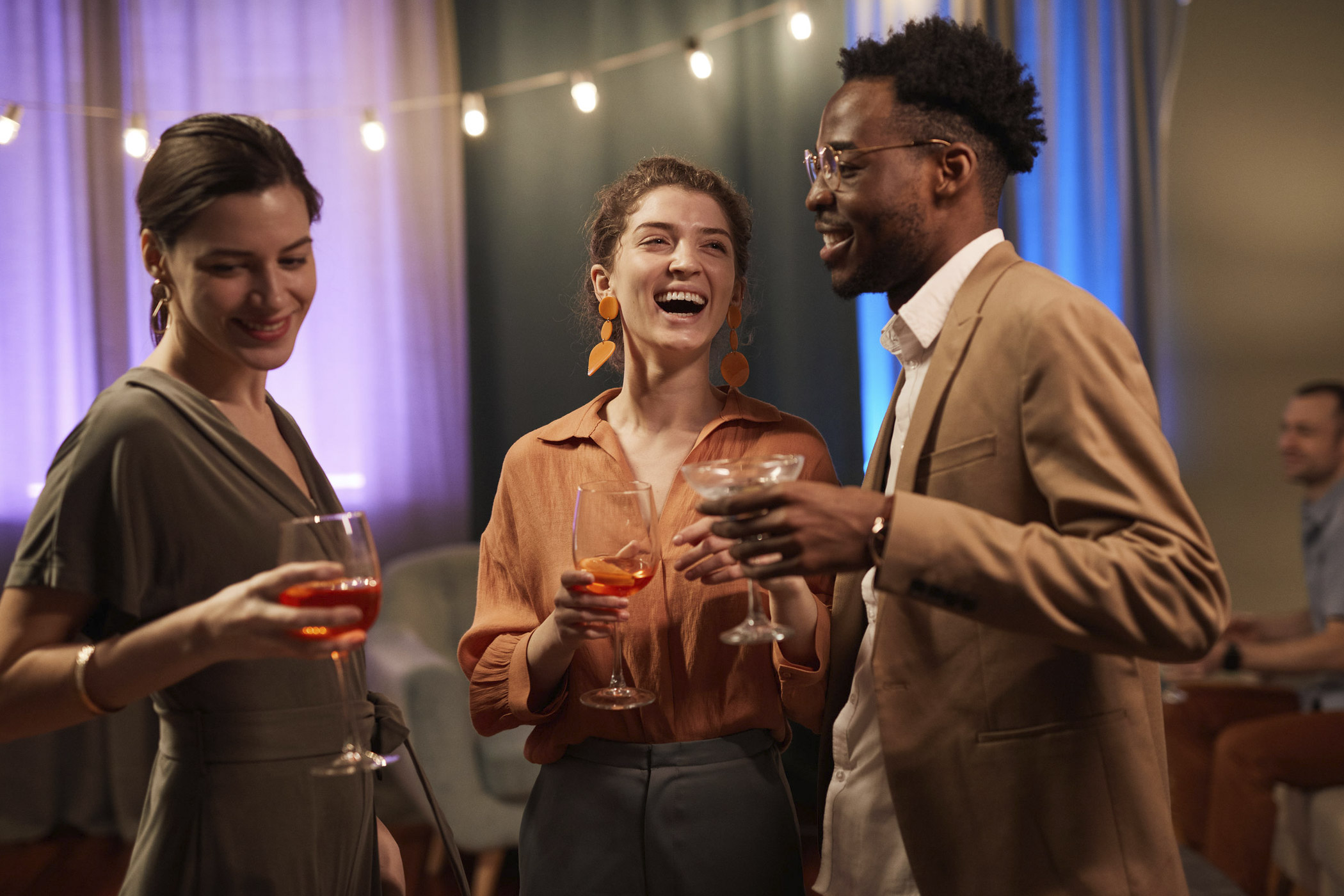 Drei Personen stehen bei einer Feier in entspannter Atmosphäre beisammen, halten Cocktail- und Weingläser in den Händen und unterhalten sich lachend. Im Hintergrund hängen Lichterketten und farbige Vorhänge sorgen für stimmungsvolle Beleuchtung.