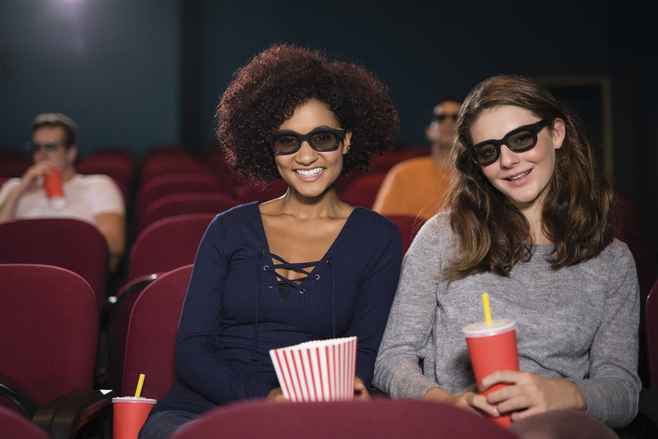 Nach Feierabend ins Kino!? / auf dem Foto: Zwei Freundinnen sitzen mit Popcorn und Getränk in einem Kino und warten auf den Beginn eines 3D-Films.