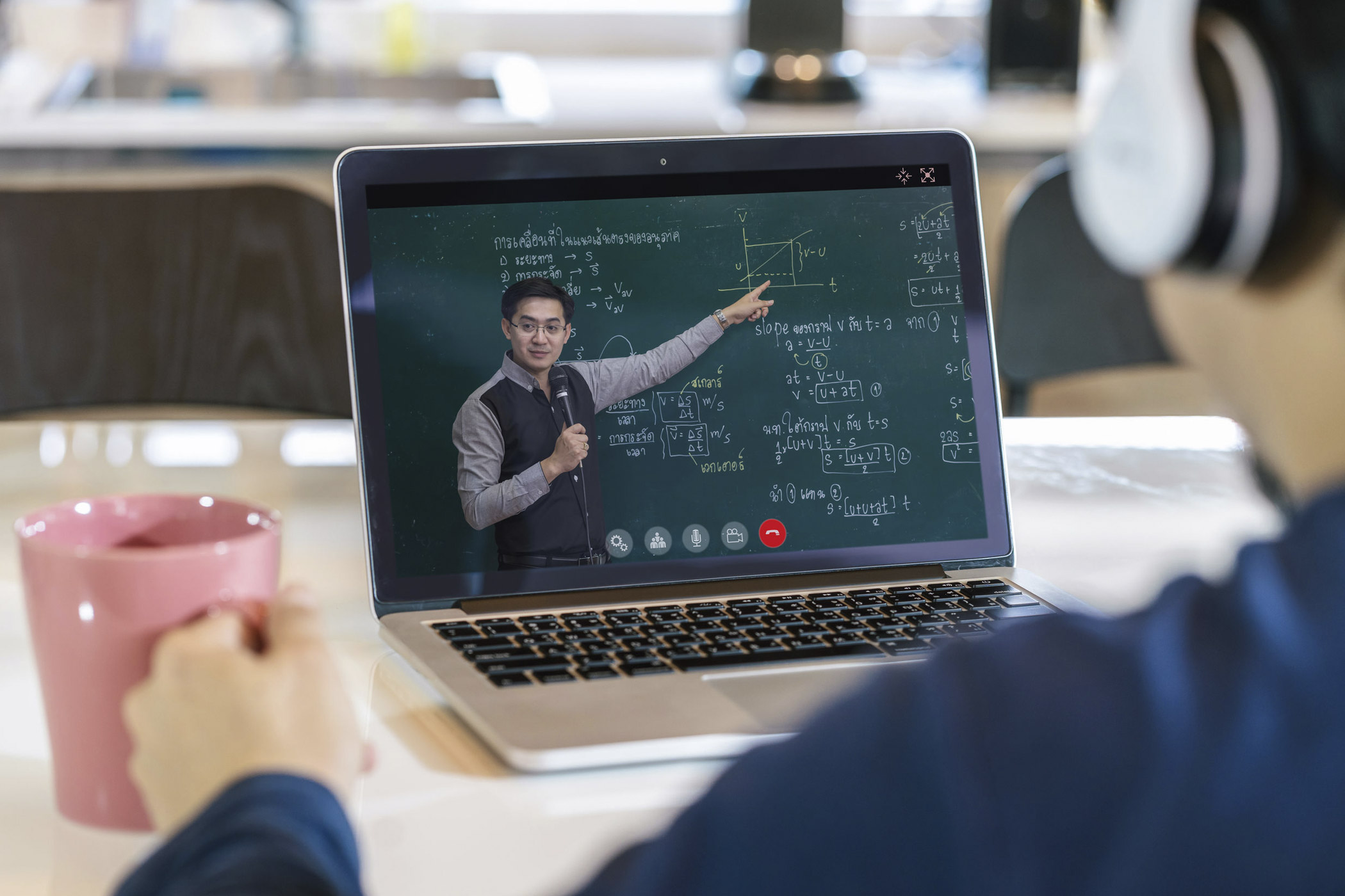 Ein Laptop zeigt eine Online-Unterrichtsstunde, bei der ein Lehrer vor einer Tafel steht und auf eine Grafik zeigt, die eine mathematische Gleichung und ein Diagramm enthält. Der Lehrer hält ein Mikrofon in der Hand und erklärt das Thema. Im Vordergrund ist eine Person zu sehen, die eine Tasse hält und auf den Bildschirm schaut, vermutlich als Teil der Online-Lernsession. Der Raum wirkt modern und gut beleuchtet.