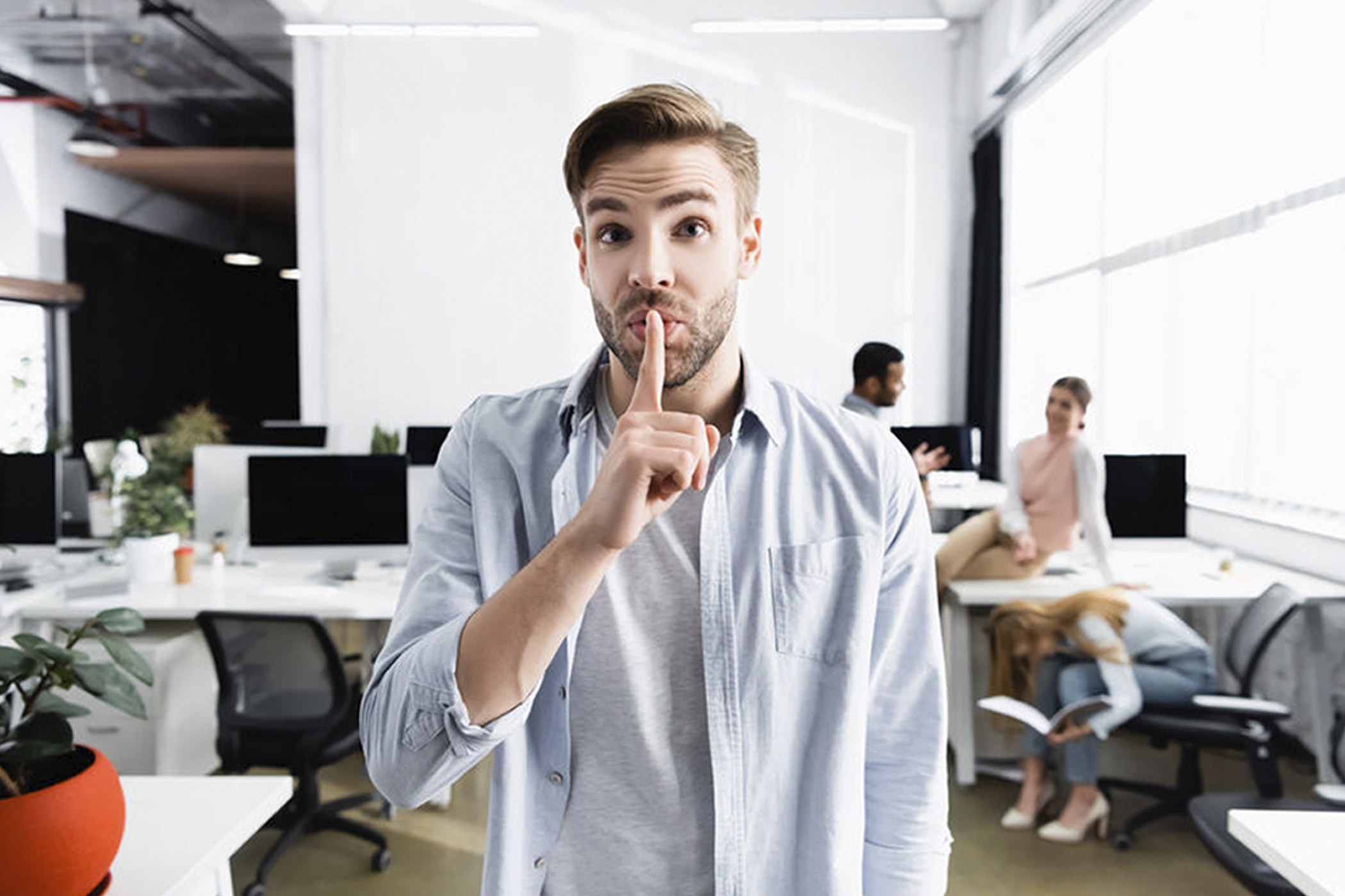 Ein junger Mann steht in einem modernen Büro und hält sich den Zeigefinger vor den Mund, um ein „Psst“-Zeichen zu machen. Im Hintergrund arbeiten mehrere Personen; eine Frau sitzt auf einem Stuhl und liest ein Buch, zwei weitere unterhalten sich locker an einem Schreibtisch. Die Atmosphäre wirkt entspannt und freundlich.