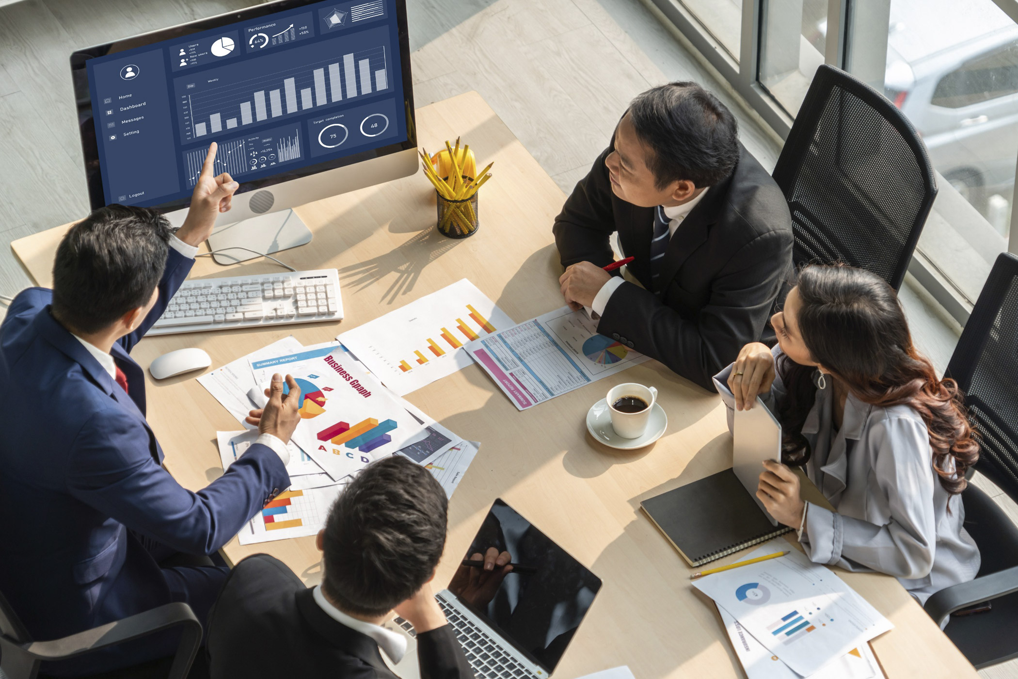 Gruppe von Geschäftsleuten in einem Meeting, die gemeinsam Diagramme und Datenanalysen auf einem Computerbildschirm und auf ausgedruckten Unterlagen besprechen.