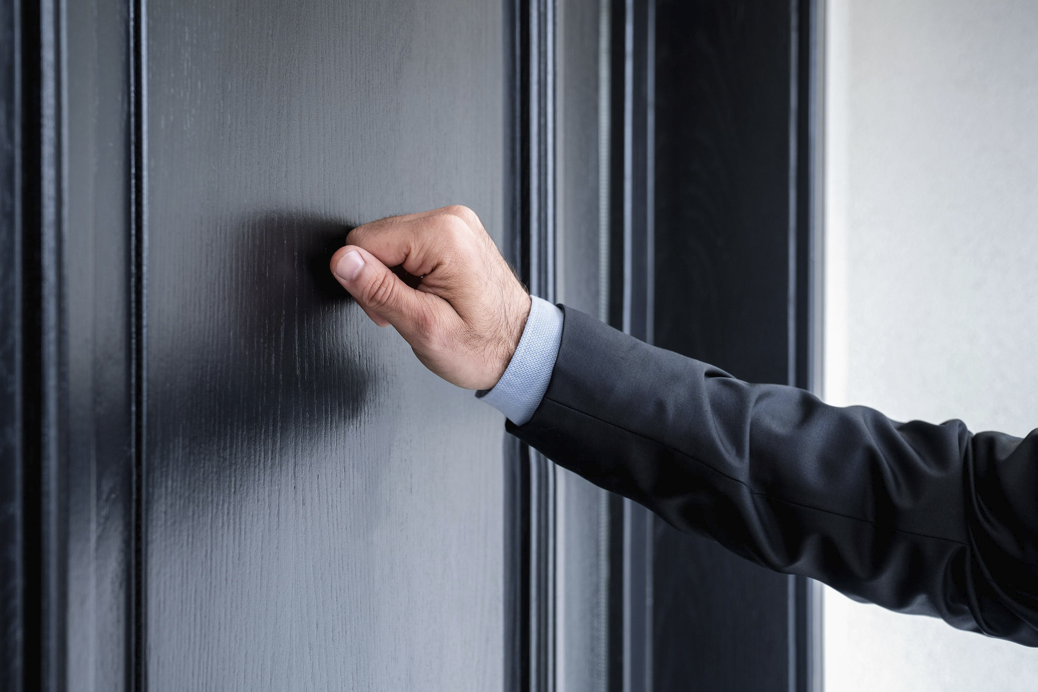Herein! Das Anklopfen ist im Arbeitsalltag ein Zeichen von Respekt. Auf dem Foto: Arm eines Mannes im Anzug, der mit den Fingerknöcheln der geschlossenen Hand an eine Bürotür klopft.