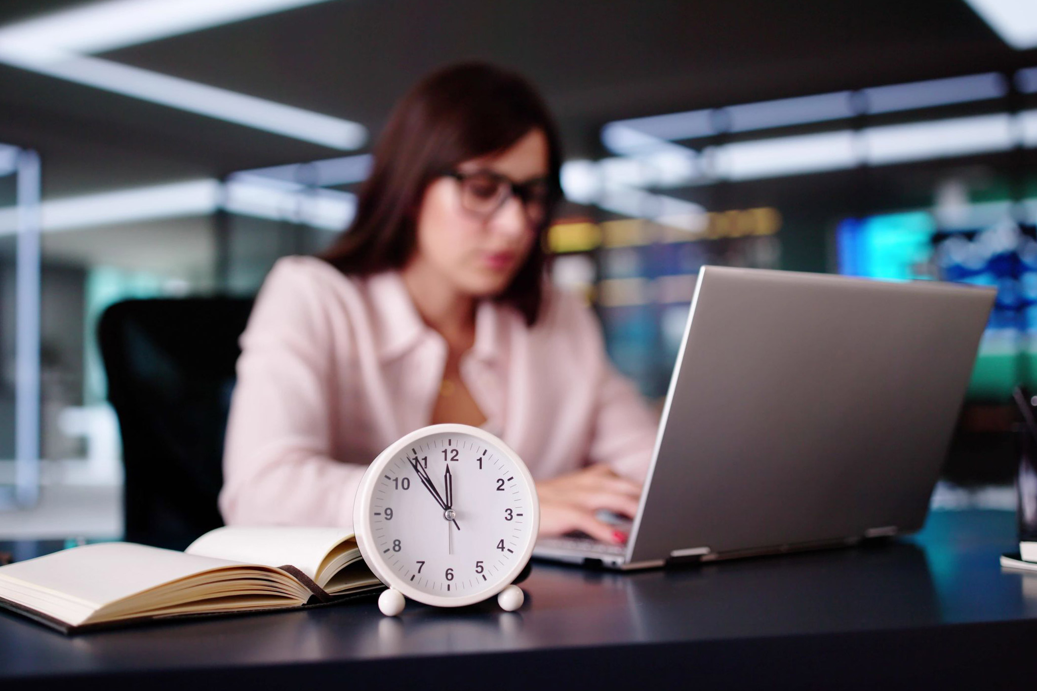 IMG Das Thema Pünktlichkeit spielt im Arbeitsalltag eine wichtige Rolle. Auf dem Foto: Eine Angestellte arbeitet am Laptop, vor ihr ein Wecker mit der Uhrzeit 11.55 Uhr